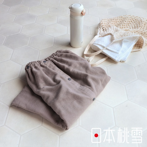 一件淺棕色棉紗混紡平口鬆緊浴裙，裙身有兩顆黑色按扣，搭配白色網狀提袋與毛巾，以及一個白色保溫瓶，整體置於六角形淺色磁磚地板上。