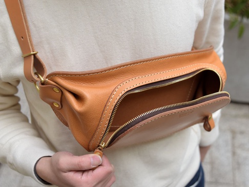 一個棕褐色皮革拉鍊斜背包，搭配黃銅拉鍊和金屬扣環，由職人手工製作，展現自然紋理和精緻縫線。