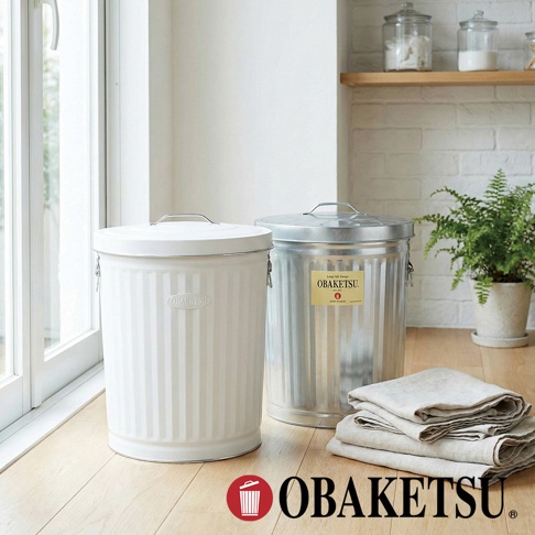 OBAKETSU 經典造型白色與銀色鍍鋅鋼製垃圾桶，擺放在木質地板上，背景有窗戶、廚房家電及綠色植物。