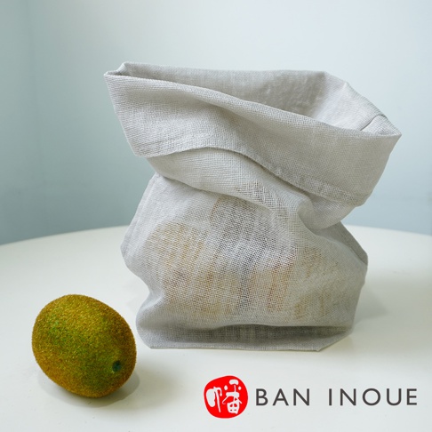 淺灰色蚊帳布材質的蔬果收納袋，袋口自然垂落，袋內裝有黃色小圓形物品，旁邊擺放一顆綠色毛絨絨的奇異果。