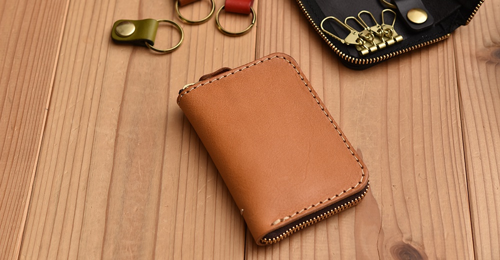 職人手作皮革多用途收納鑰匙包，淺棕色皮革，附有縫線細節與拉鍊。背景為木質桌面，另有黑色鑰匙包及鑰匙圈。