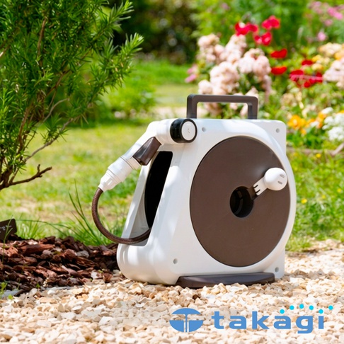 一款簡約現代的takagi白色園藝澆花灑水槍組放置在花園碎石地上，主體為方形收納盒設計，搭配深褐色轉盤與頂部提把，側邊連結著噴水槍頭，背景呈現綠意盎然的戶外庭園景致。