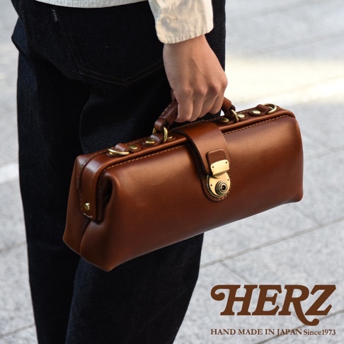 HERZ 品牌標誌與 HAND MADE IN JAPAN Since1973 字樣，以及一個由皮革製成的棕色長方形手提包，包包上配有金色扣環和金屬配件。