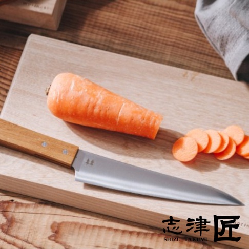 切菜刀放在木砧板上，旁邊放著一根胡蘿蔔和幾片切好的胡蘿蔔，刀柄上有志津匠的標誌。