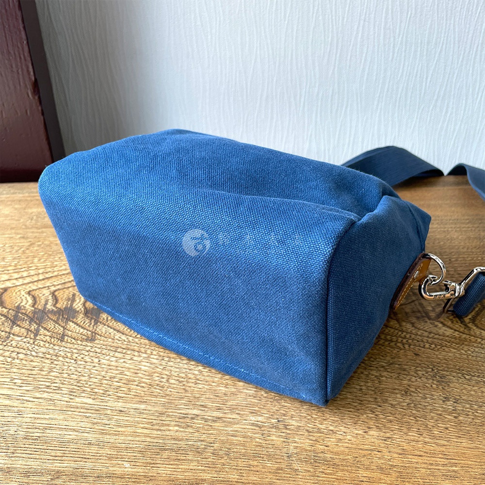 深藍色石洗帆布散步包，側面有皮革裝飾與金屬掛鉤，放置於木紋桌面上。