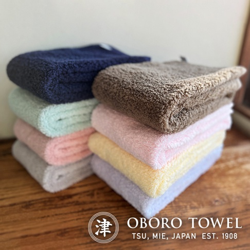 堆疊的毛巾展示了多種柔和色彩，包括海軍藍、淺綠、粉紅、灰色、棕色、淺黃和淺紫色。毛巾材質看起來蓬鬆柔軟，觸感細膩。