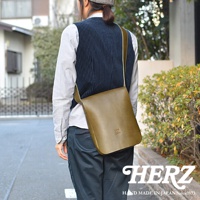 一名男子身背日本 HERZ 品牌皮革軟皮直式斜背包 CK-98，該背包為大地色，呈現職人手作的質感。