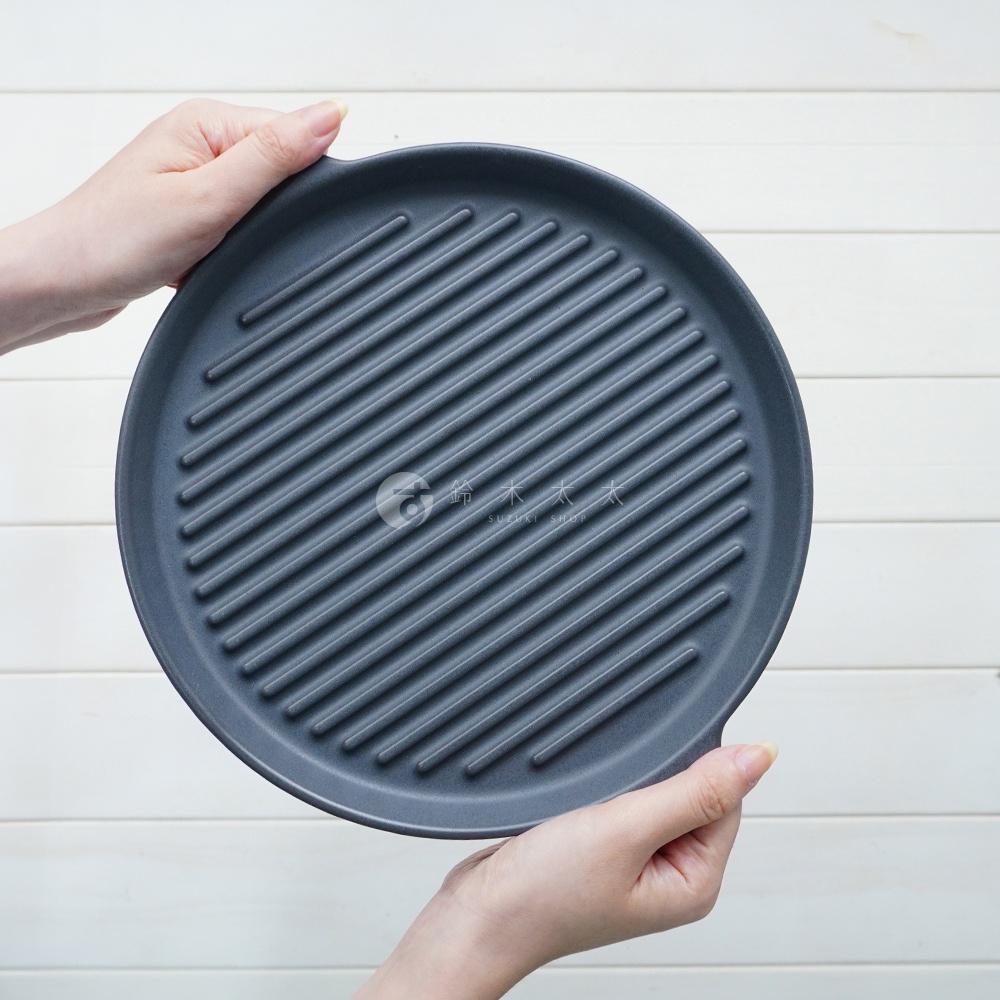 兩隻手捧著一個深灰色、圓形、帶有平行波浪紋路的陶製烤盤。