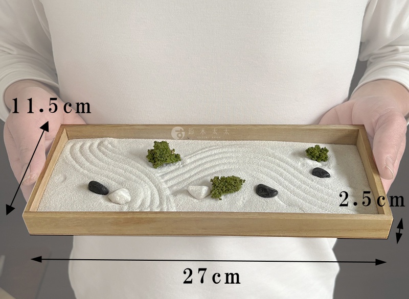這張照片展示了一款長方形的桌面枯山水擺飾，由一人手持呈現。木製邊框的容器內裝滿白沙，沙面上繪有波浪紋路，並點綴著幾顆黑色與白色的石子以及幾簇綠色苔蘚裝飾，整體風格簡約禪意。圖片標註了模型尺寸，寬度為二十七公分，深度為十一點五公分，高度為二點五公分。