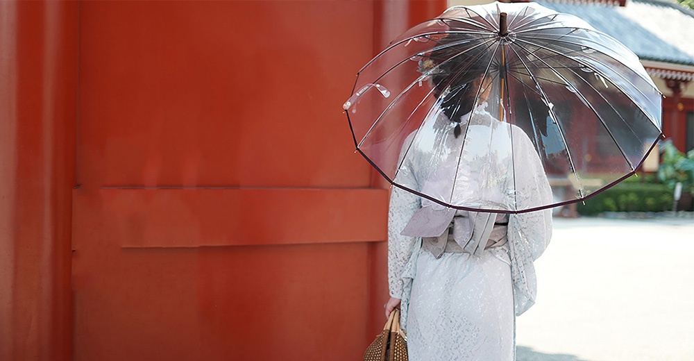 一名女子撐著透明傘，傘面有細緻的櫻花圖案，傘骨與提把為樺木材質，女子身著淺色和服，站在紅色背景前。