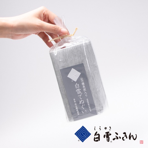 手持一條以透明塑膠袋包裝的灰色紗布長巾，袋口以麻繩綑綁。長巾表面可見細緻的織紋，袋上貼有深灰色標籤，印有白色日文與圖案，產品名稱為「紀州備長炭紗布長巾」。