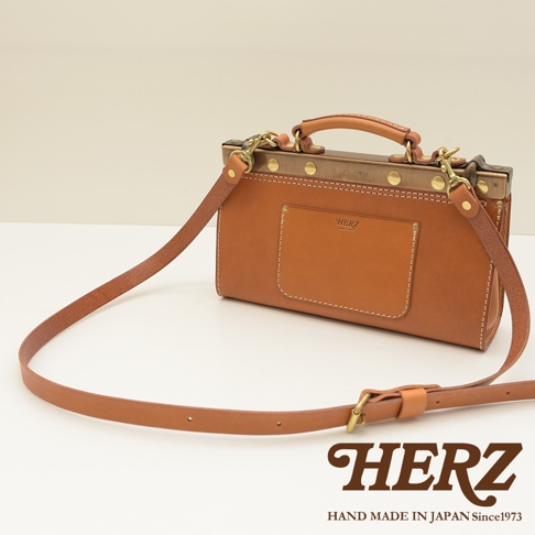 一只棕褐色皮革製作、復古風格的兩用手提包，包身帶有方形外袋、品牌壓紋及縫線細節，搭配金屬口金、銅色铆釘、提把及可拆卸斜背帶，展現職人手工質感。