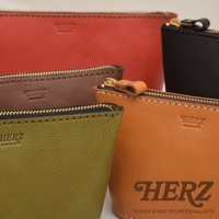 職人手作皮革三角收納包，展示多款顏色，包括紅色、黑色、棕色、綠色和橘色。包款採用皮革材質，搭配金屬拉鍊，並印有品牌標誌。