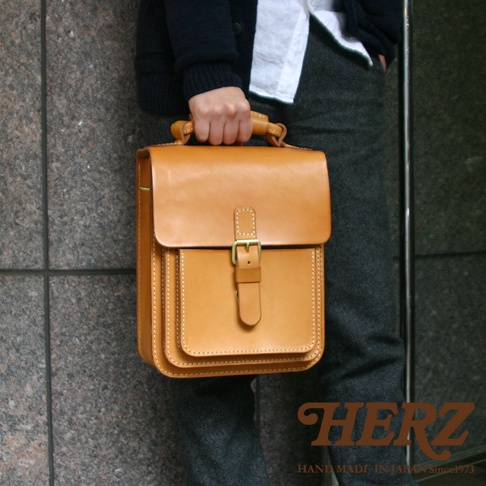 職人手作的駝色皮革單肩兩用小方包，包身結構分明，正面有帶金屬釦的翻蓋與置物袋，車縫線清晰。