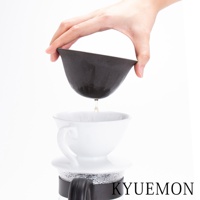 一隻手持深灰色、帶有紋理的陶瓷濾杯，杯中滴出液體，下方置有白色陶瓷咖啡杯，杯座為玻璃材質，整體置於白色背景上。