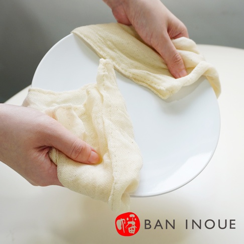 一雙手正在使用兩塊米白色、紋理明顯的有機蚊帳棉織廚房拭巾擦拭一個白色圓盤。