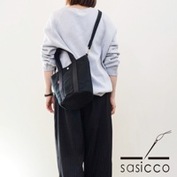 一位女性身著淺灰色衛衣和黑色條紋長褲，側背一個黑色三河木綿刺子織兩用水桶包。包包為圓筒狀，有兩個短提把和一條斜背帶。