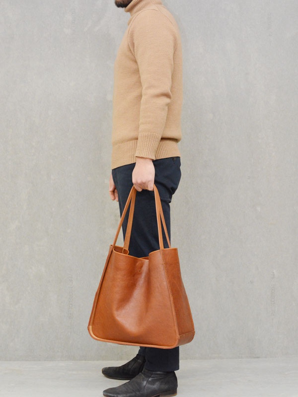 一位男士手持一個棕色皮革方形托特包，包包側面有縫線裝飾，提把為皮革材質。