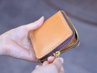 一隻手拿著一個棕褐色皮革製作的短夾，拉鍊部分有深棕色布邊與金色拉鍊頭，縫線為白色。