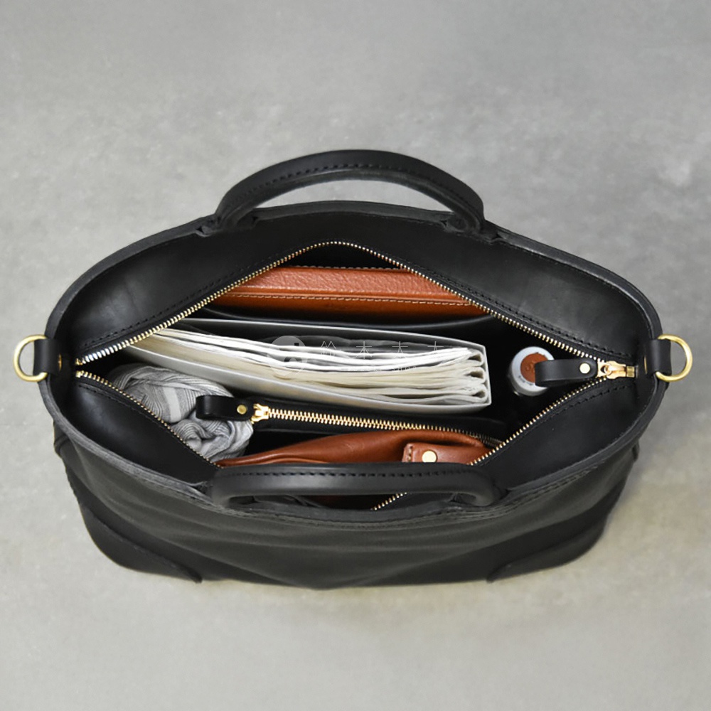 一個敞開的黑色皮革托特包，裡面裝有文件、錢包和一個小型圓柱形物品，側邊有提把和可拆卸的肩帶。