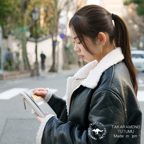 一位年輕女子身穿黑色皮夾克，手裡拿著灰色長夾，夾克內襯為白色絨毛，背景模糊，街景中有人影及建築物，右下角有 TUTUMU 的 Logo 和文字 TAKARAMONO TUTUMU Made in jpn。