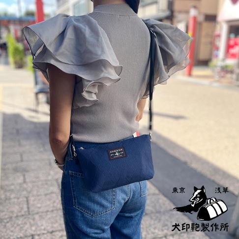 深藍色細條紋斜背包，配有黑色肩帶和銀色金屬拉鍊。包上繡有品牌標誌。