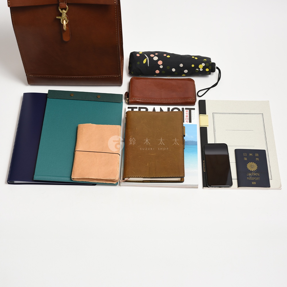 深棕色皮革紙袋風後背包，搭配黑色印花摺疊傘、棕色皮革長夾、深藍色筆記本、綠色文件夾、大地色系筆記本，以及手機、日本護照。