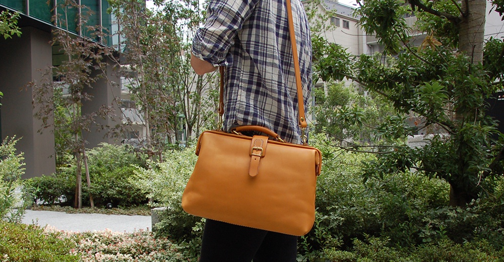 一款淺棕色皮革軟式杜勒斯包，配有可調節的肩帶和金色搭扣，由一位身穿格紋襯衫的人士攜帶，背景是綠色植物和建築物。