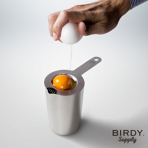 一只手正在將雞蛋打入一個銀色的不鏽鋼杯中，杯內有一個蛋黃和部分蛋白。杯子帶有一個短柄，上面印有「BIRDY. SUPPLY」的標誌。