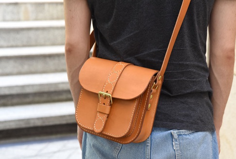 棕褐色皮革製成的單肩包，配有黃銅扣和十字繡裝飾的翻蓋，以及裝飾性的縫線。