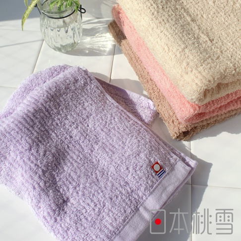 一條紫色細絨毛巾，搭配一疊米色、粉紅色和淺棕色毛巾，皆為柔軟材質，帶有細緻紋理，角落有品牌標誌。