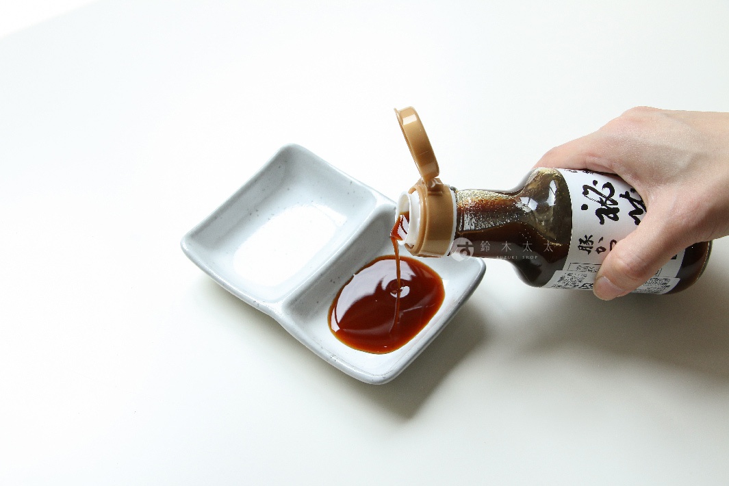 一只手拿著標有日文的瓶子，將深棕色的醬汁倒入一個淺灰色、有兩個隔間的陶瓷碟中。