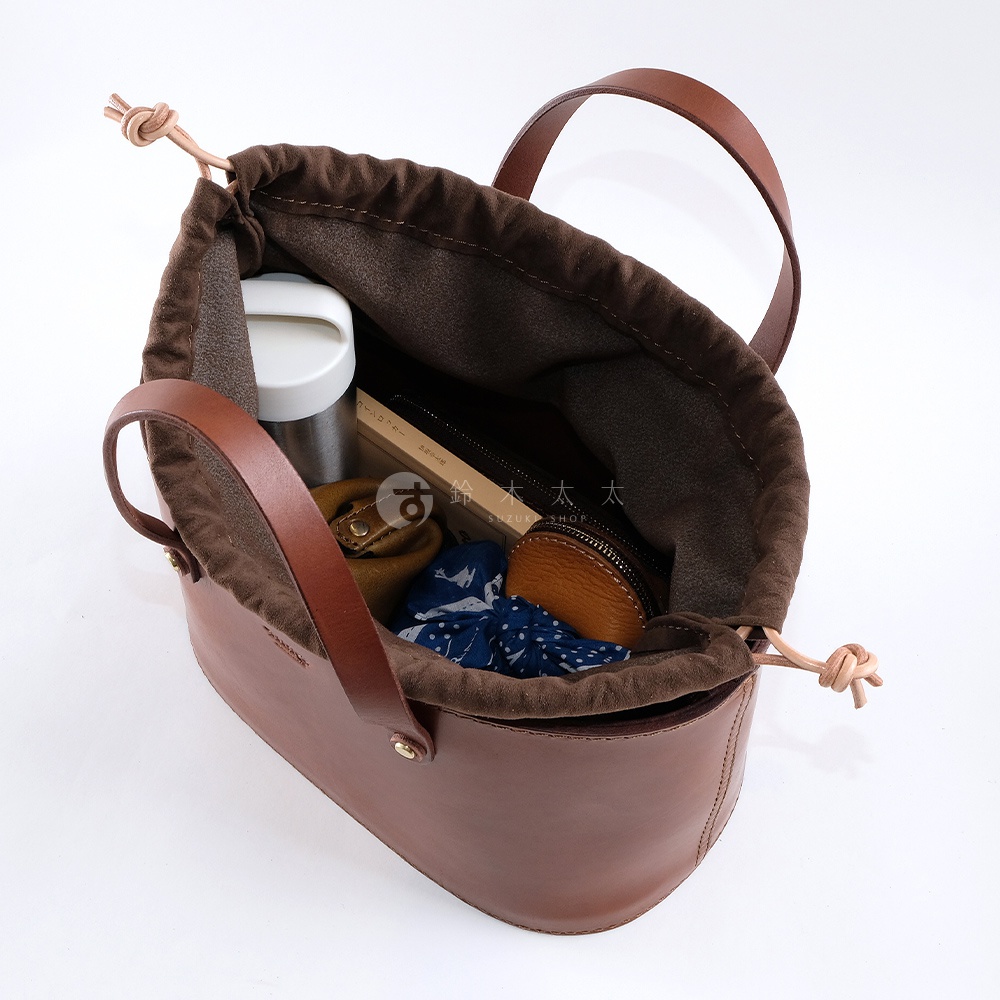 職人手作皮革束口手提包，咖啡色牛皮材質，內襯為深棕色帆布，束口處以皮革繩繫結。包內置有保溫杯、筆、小皮夾及印花頭巾。