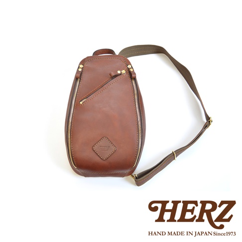 職人手作皮革多功能收納斜背包，採用光滑棕色皮革製成，配有金色拉鍊和可調節的織帶肩帶。包身呈水滴狀，設有一個斜向拉鍊前袋和一個菱形皮革標誌。