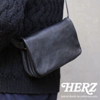 黑色皮革斜背包，帶有翻蓋和金色金屬扣，斜挎於穿著黑色針織衫的人身上。