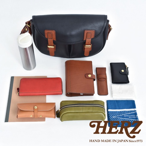 黑色皮革斜背包，搭配棕色皮革飾邊與帶扣。周圍擺放著紅色長夾、棕色筆記本、棕色筆套、黑色零錢包、軍綠色化妝包、藍色印花手帕，以及一個銀色保溫瓶。