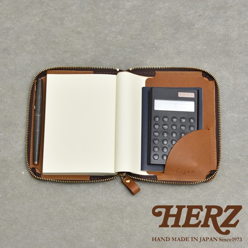 職人手作皮革Organ記事本收納包，打開狀態，內有空白筆記本、黑色計算機及一支筆，收納包為棕色皮革材質，拉鍊為咖啡色。