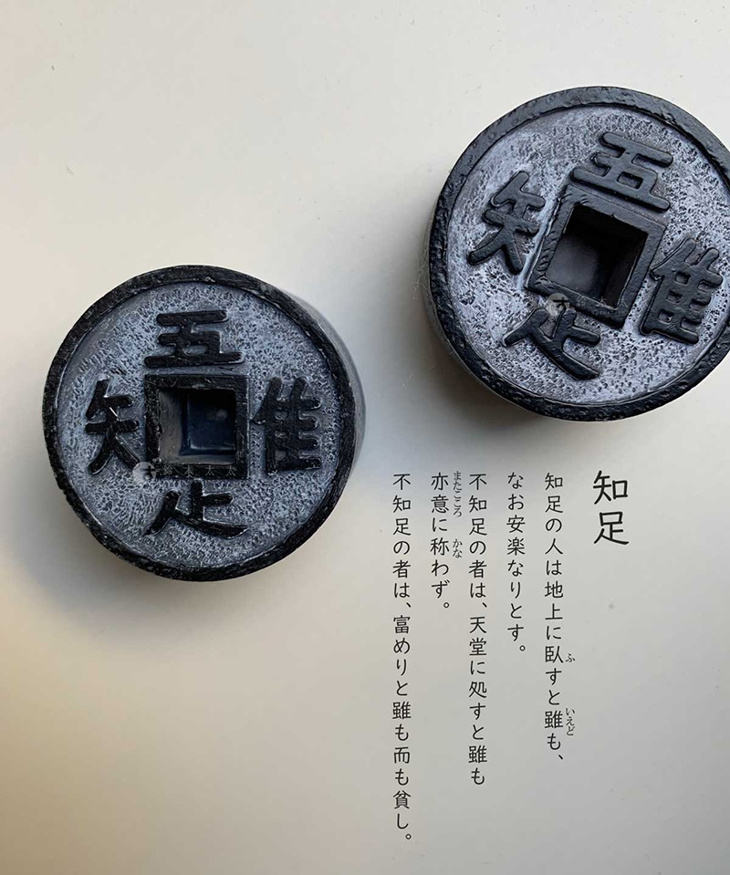 圖片文字：知足。知足の人は地上に臥すと雖も、なお安楽なりとす。不知足の者は、天堂に処すと雖もまた意に称わず。不知足の者は、富めりと雖も而も貧し。兩枚圓形仿古錢幣模型排列於白色背景上，錢幣中心為方形孔，表面刻有吾、唯、知、足四個漢字，錢幣質地呈現粗糙的金屬感或石材感，右側配有與知足哲理相關的日文說明文字。