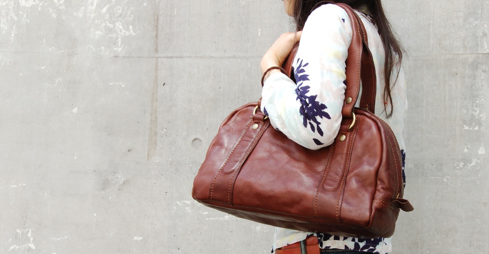 咖啡色水洗皮革迷你波士頓包，配有雙提把和金屬拉鍊，由一位女士夾在腋下，身穿印花上衣。