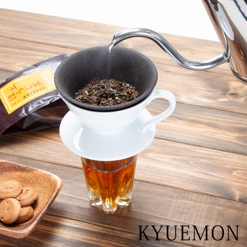 白色陶瓷濾杯組，內含黑色濾網，上方盛有茶葉，正由金屬細嘴茶壺注入熱水。濾杯組下方置於透明玻璃杯中，杯內有琥珀色茶湯。左側木盤上有數枚餅乾，後方為一包黃色標籤的茶葉包。