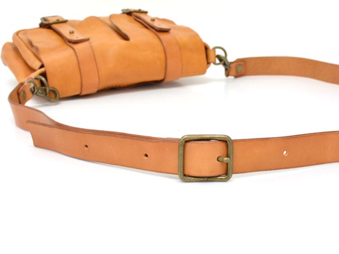 淺棕色皮革製雙口袋肩背包，有兩個外部口袋，以金屬扣和皮帶固定，搭配一個可調節的皮革肩帶，肩帶上有方形金屬扣。