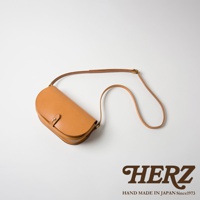 職人手作皮革迷你馬鞍包，採用天然皮革材質，呈現溫潤的焦糖色。包款設計為半月形，配有可調式長背帶和金屬鈕扣開合。