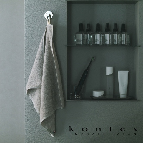 灰色牆面掛著一條淺米色的 KONTEX 棉質方巾，右側置物架上整齊擺放著多瓶透明護膚液、電動牙刷及白色保養軟管，整體氛圍呈現出簡約質感的衛浴生活空間。