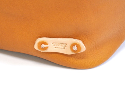 焦糖色皮革托特包的特寫，展示了品牌標誌「HERZ」和「MADE IN JAPAN」的壓印，以及固定細節。