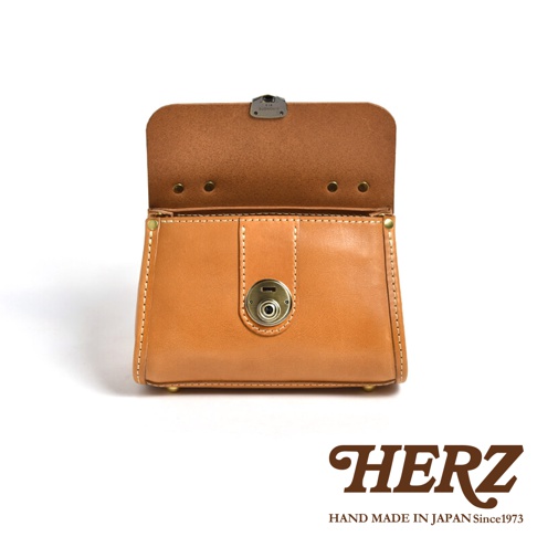 職人手作皮革迷你手提包，採用天然皮革製作，呈現溫潤的焦糖棕色。包款正面有一個金屬旋轉鎖扣，掀蓋式設計，縫線精緻，整體風格簡約而復古。