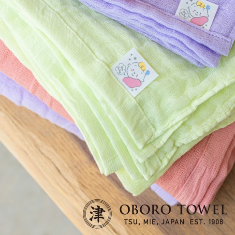 三層紗純棉寶寶裹身毯，包含淺綠、淡紫、珊瑚粉三色，質地輕柔，帶有可愛寶寶圖案的品牌標籤。