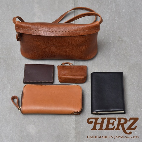 HERZ品牌標誌，下方有HAND MADE IN JAPAN Since1973字樣，圖片展示了多款皮革製品，包含一個棕色斜背包、一個深棕色皮夾、一個棕色零錢包、一個淺棕色長夾以及一本黑色筆記本。