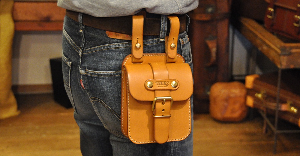 淺棕色皮革製成的經典腰袋，透過縫線固定，袋蓋處有皮帶與金屬釦環，可掛於腰帶上，適合攜帶手機等隨身物品。