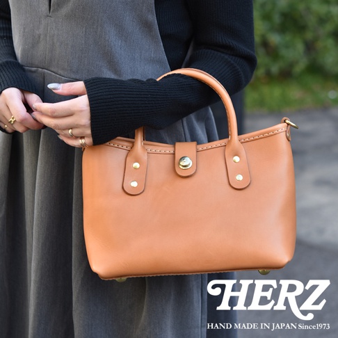 職人手作皮革迷你托特包，採用光滑棕色皮革，配有金色金屬扣和鉚釘。包包有兩個短提把，並透過車縫線裝飾，整體呈現簡約實用的風格。