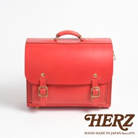 一個鮮紅色的皮革橫式書包，具有頂部提把、兩個前方的皮帶扣和金屬腳釘。書包的右下方印有HERZ字樣和HAND MADE IN JAPAN Since1973的字樣。